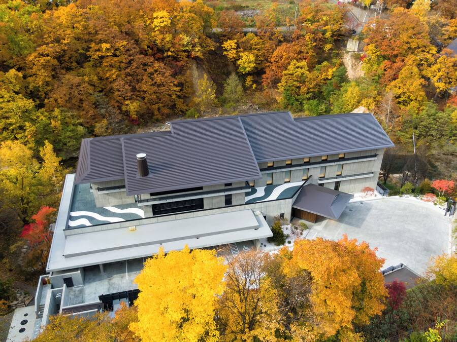 A modern building with a flat roof and a large parking area surrounded by autumn-colored trees.