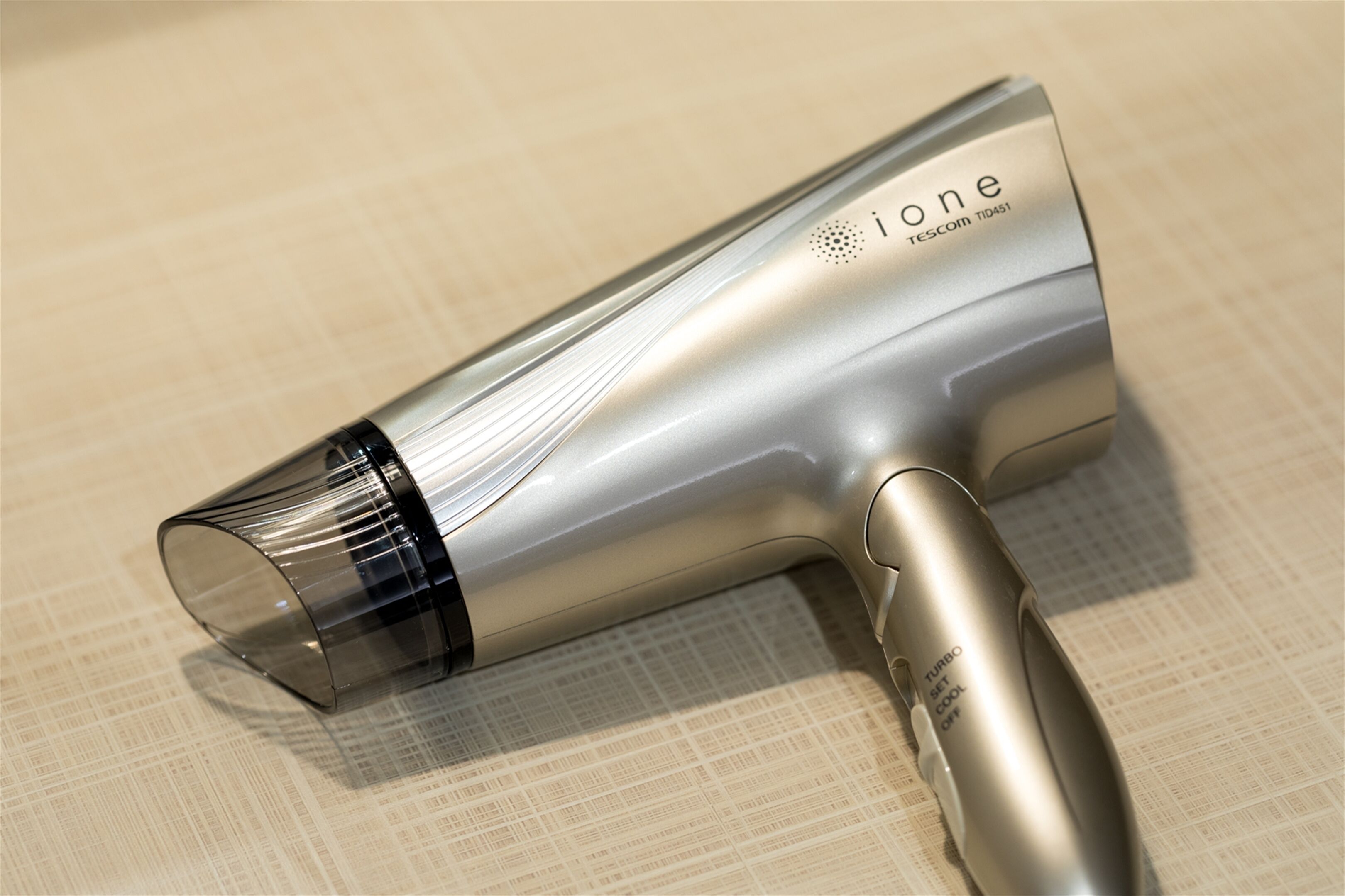 A metallic hairdryer with a handle and a cord, placed on a textured surface.