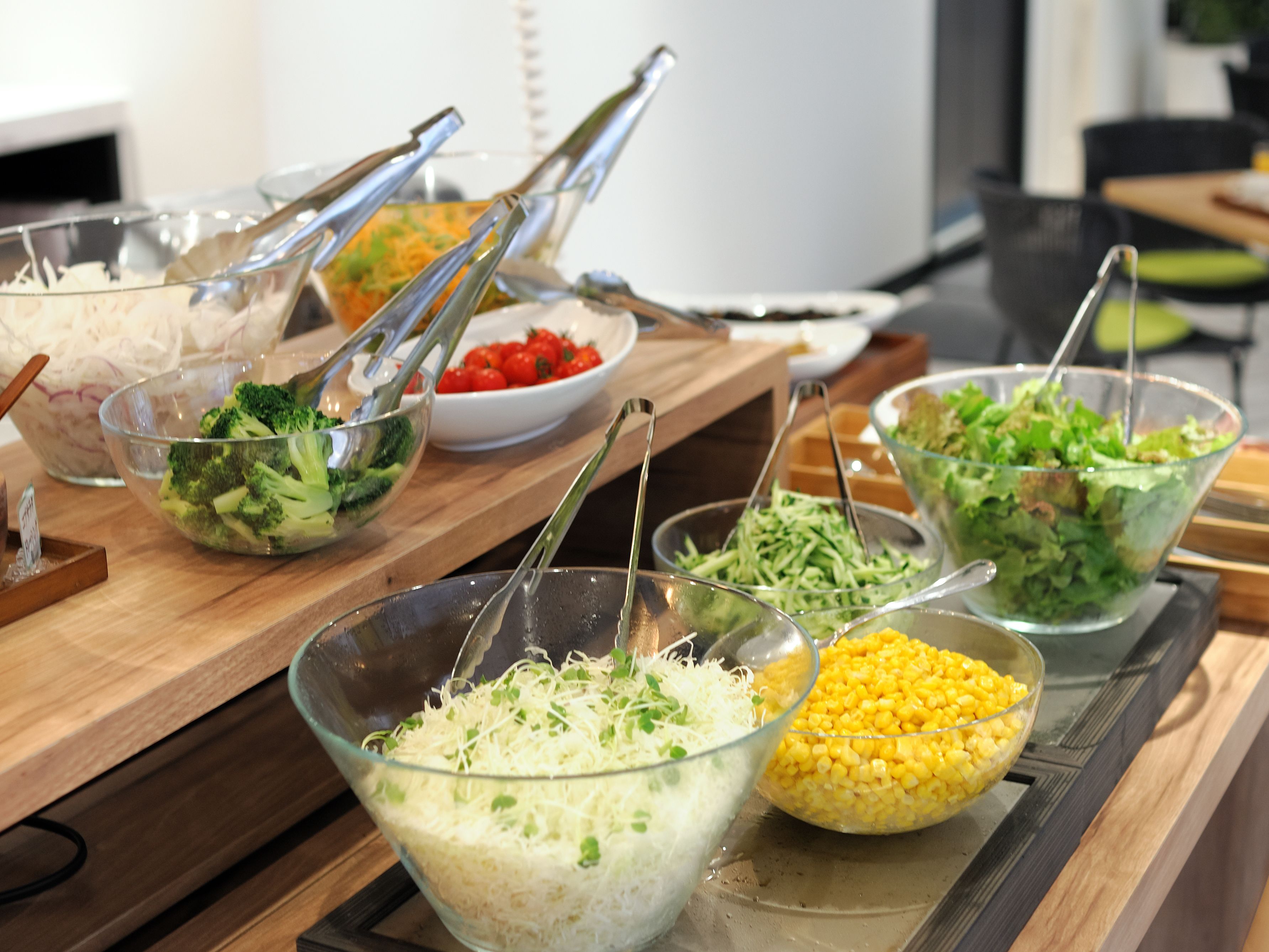Una mesa de buffet con diversos alimentos en recipientes transparentes, como brócoli, tomates, queso rallado, maíz y una ensalada verde.