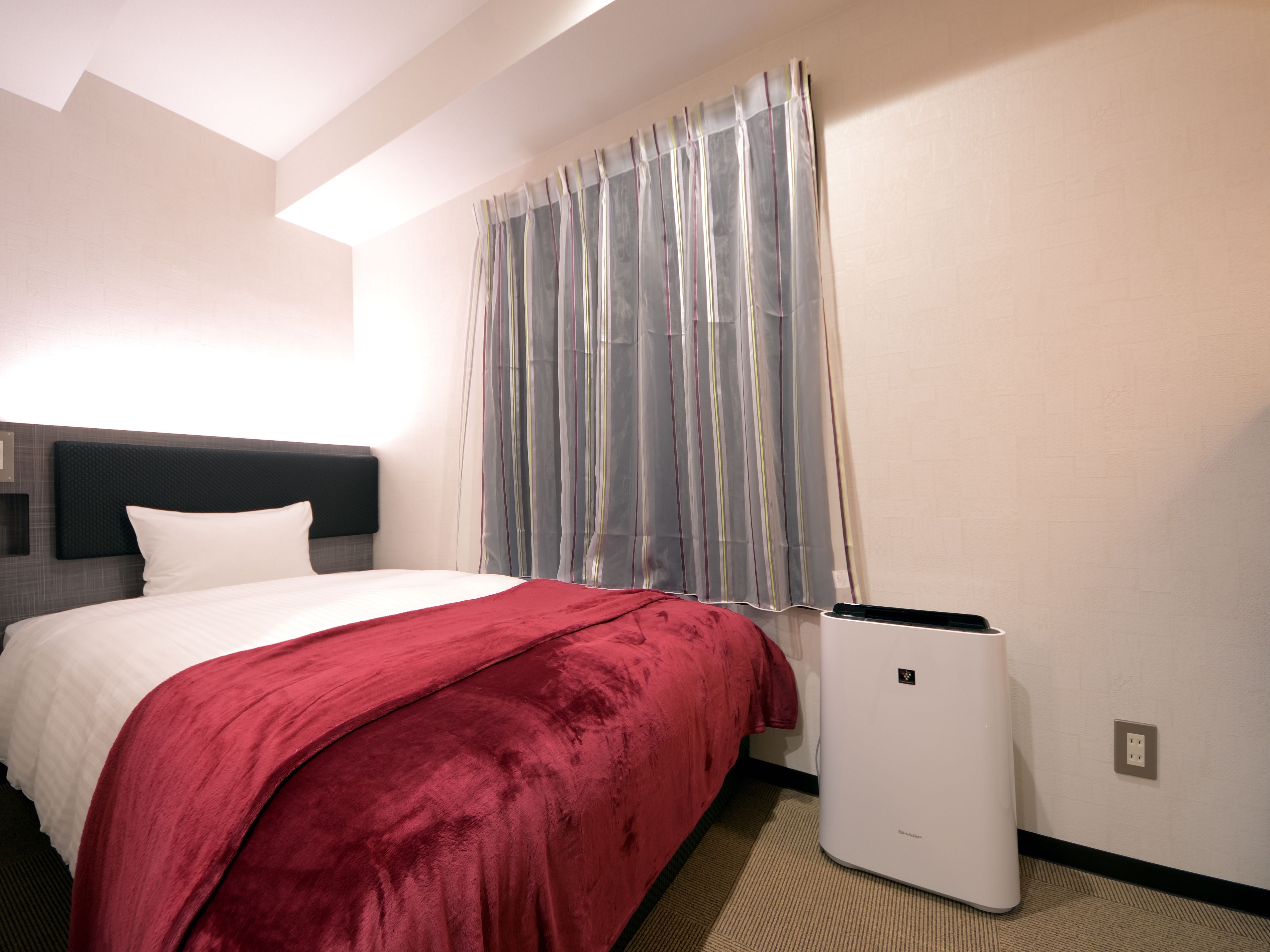 A hotel room with a bed, a headboard, a window with curtains, and a wall-mounted heater.