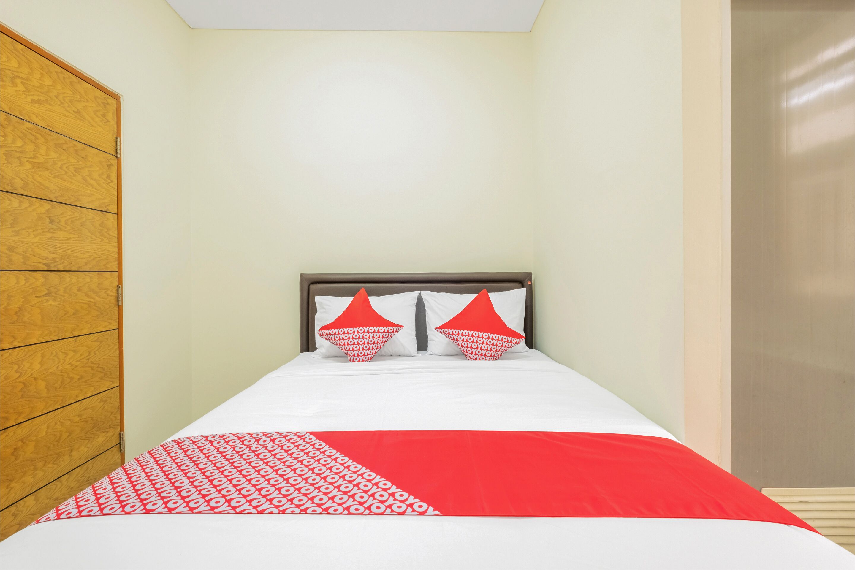 A single bed with a white and red bedspread, two red pillows, and a wooden wardrobe to the left.