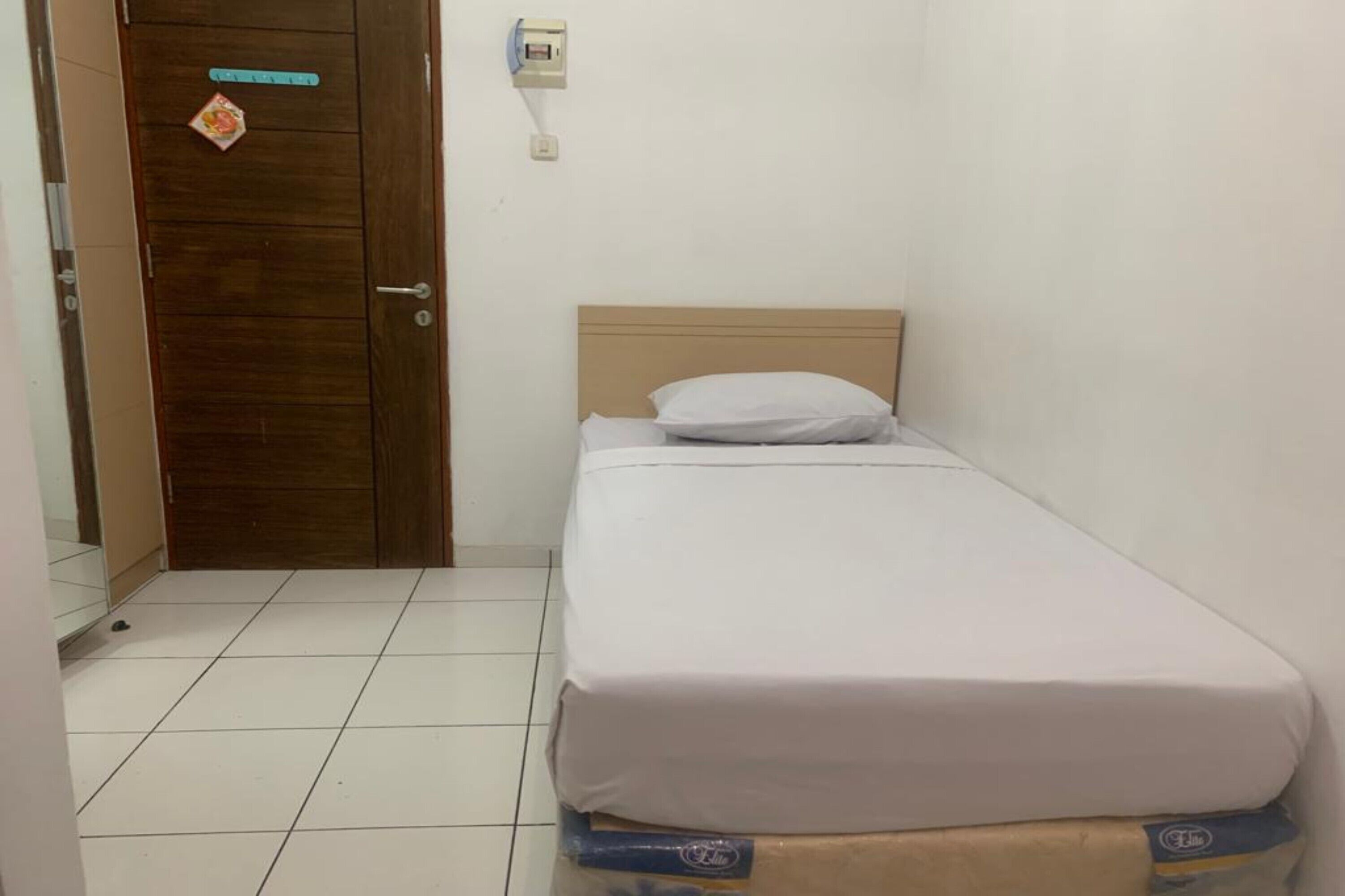 Une petite chambre avec un lit simple, une porte en bois, une table de chevet et un interrupteur fixé au mur.