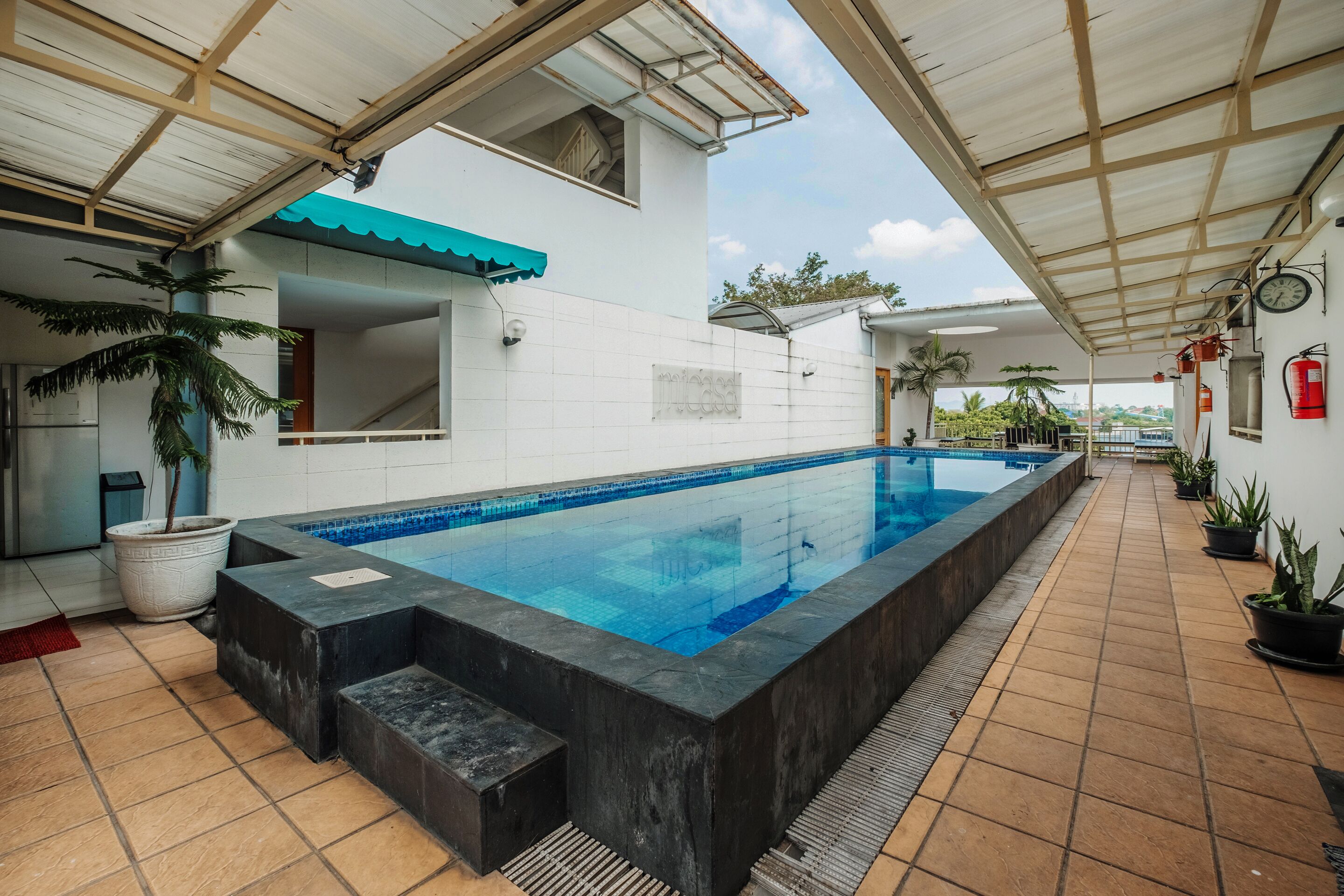Une piscine avec une terrasse carrelée, un patio couvert et des plantes en pot.
