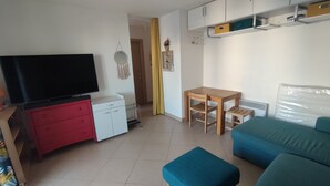 Una habitació petita i ben il·luminada amb un televisor sobre una petita còmoda vermella, una taula de fusta amb cadires, un sofà blau i un armari blanc amb prestatgeries.