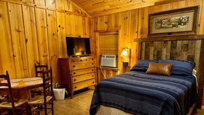 A wooden-paneled room with a bed, a television, a dresser, and a table with chairs. - Cabin at Sunset Ranch with Sunset Resort on Medina Lake access! (Mico)