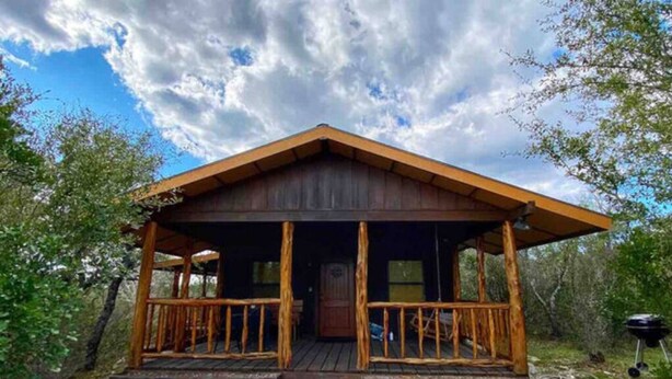 A rustic cabin with a wooden porch and a door, surrounded by trees. - Cabin at Sunset Ranch with Sunset Resort on Medina Lake access! (Mico)