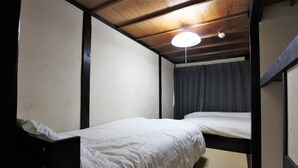 A small, dimly lit room with two single beds, wooden beams on the ceiling, and a plain curtain on the window. - The Neighbors Tsukamoto (Osaka)