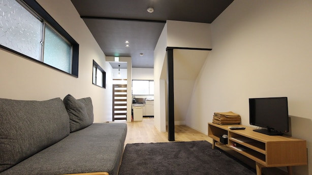 A modern living room with a grey sofa, a TV on a wooden stand, and a kitchen in the background. - The Neighbors Tsukamoto (Osaka)