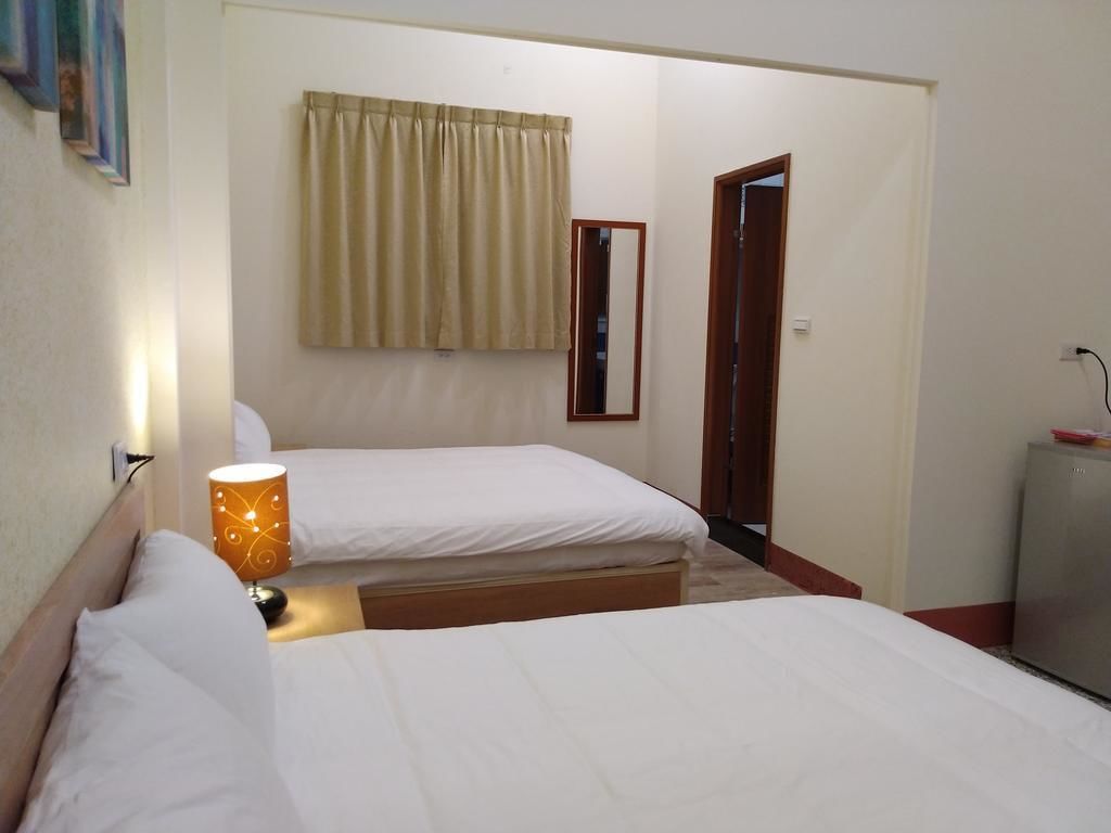 Une chambre d’hôtel avec deux lits, un miroir, une fenêtre avec des rideaux, une lampe et un petit meuble.