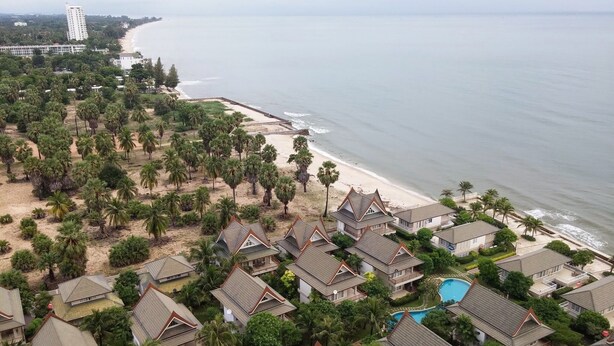 Un resort costero con villas tipo cabaña, una playa y un gran hotel al fondo.