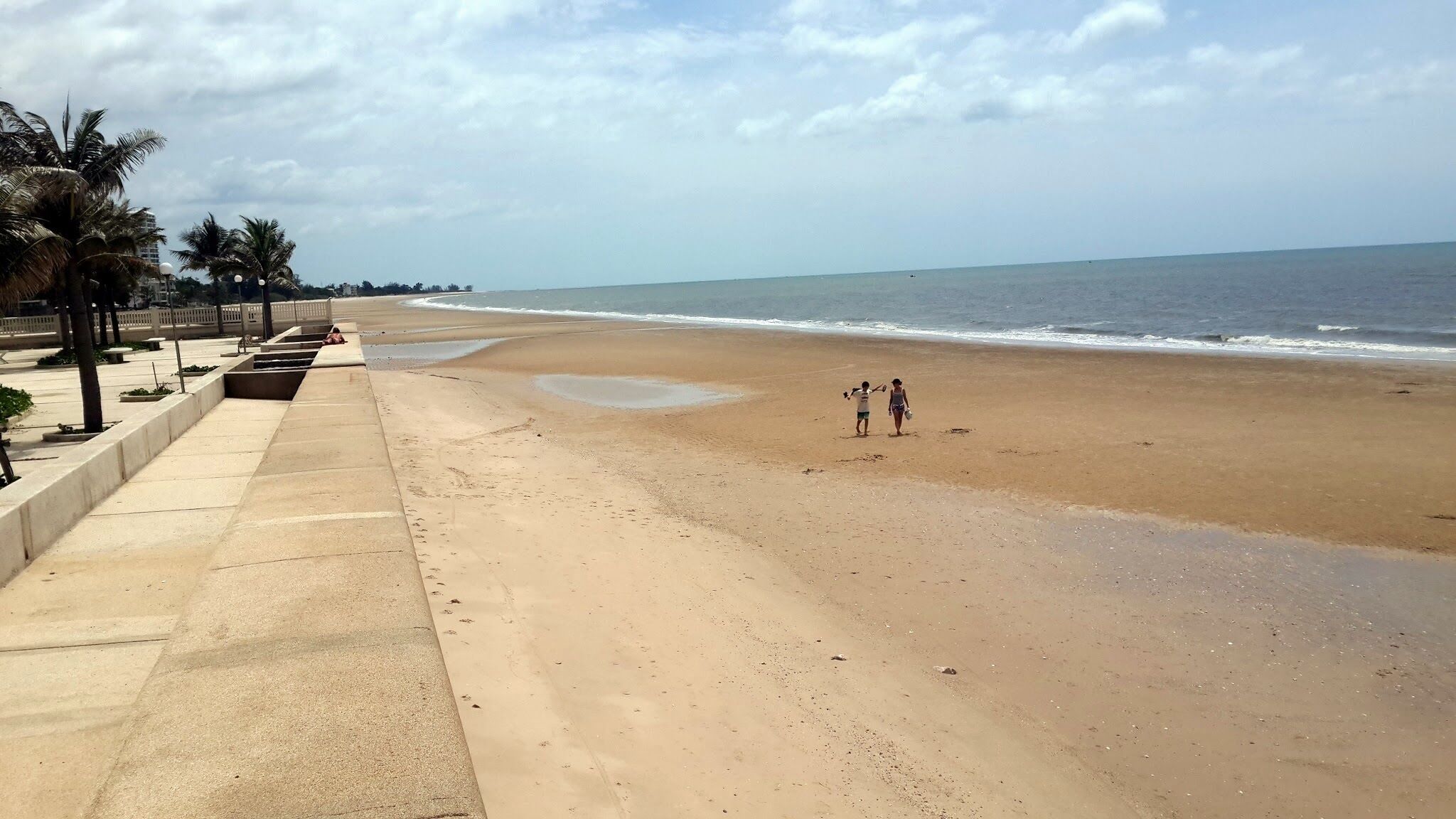A wide sandy beach with a concrete walkway on the left, palm trees, and a calm sea.