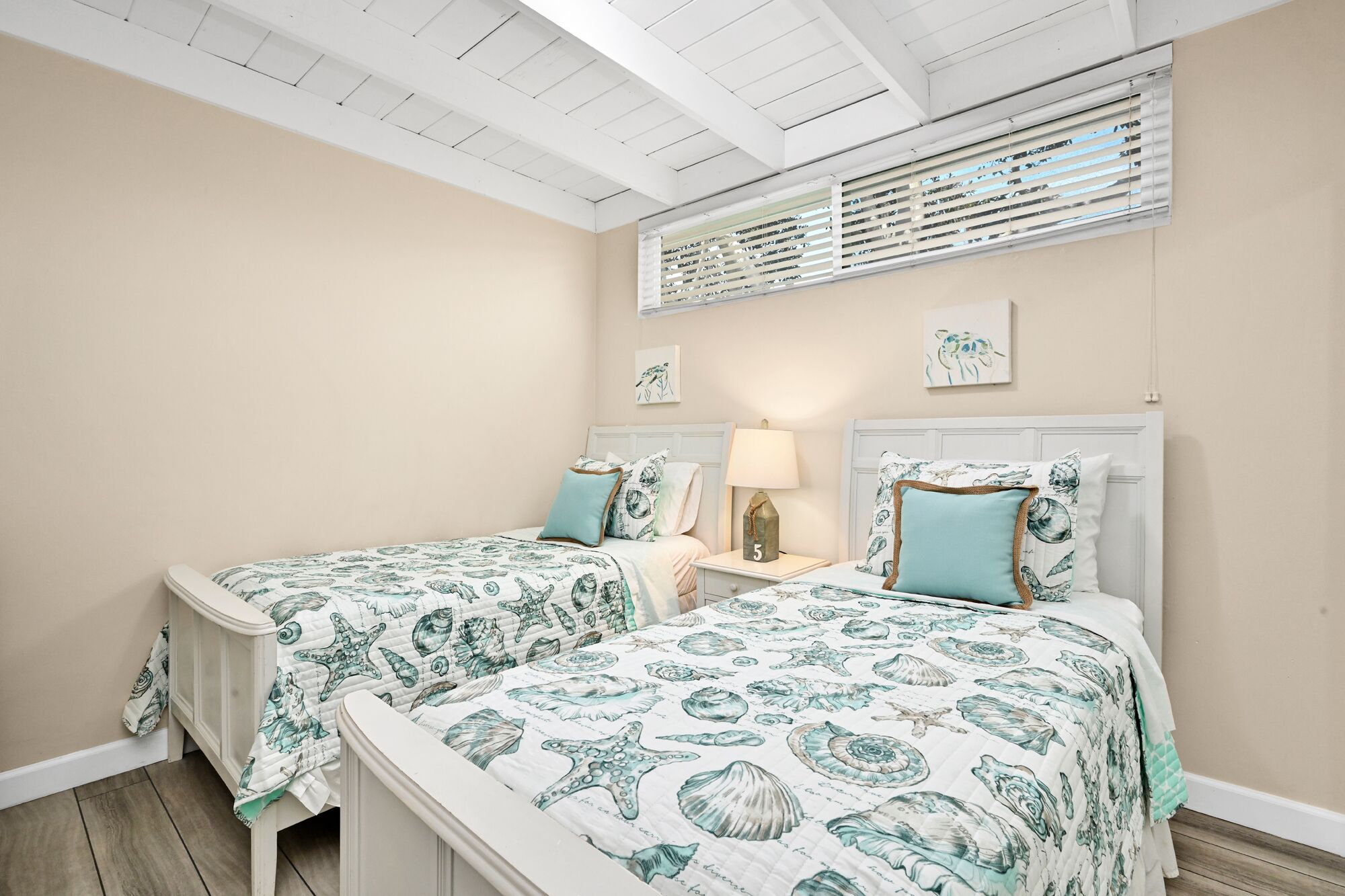 Een slaapkamer met twee bedden, elk met passend beddengoed met afbeeldingen van zeedieren. Tussen de bedden staat een nachtkastje met een lamp en een klein decoratief item. De kamer heeft een schuine plafond met houten balken en een raam met zonwering.