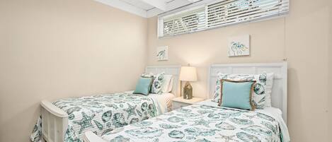 Een slaapkamer met twee bedden, elk met passend beddengoed met afbeeldingen van zeedieren. Tussen de bedden staat een nachtkastje met een lamp en een klein decoratief item. De kamer heeft een schuine plafond met houten balken en een raam met zonwering.