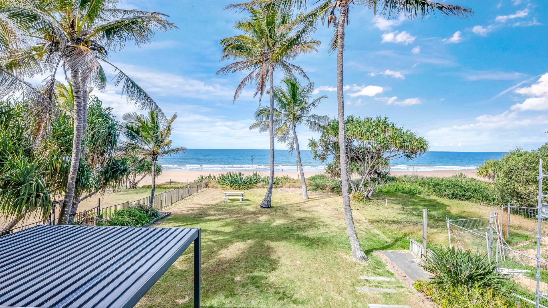 A coastal property with a grassy lawn, palm trees, a metal fence, and a view of the beach and ocean.