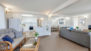 A modern living room with a grey sofa, a wicker chair, a glass coffee table, and a kitchen in the background. - Horizons 3-16 Miller Bargara (BARGARA)