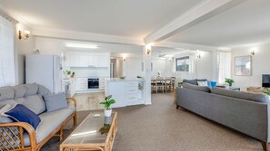 Una sala de estar moderna con un sofá gris, un sillón de mimbre, una mesa de centro de vidrio y una cocina al fondo.