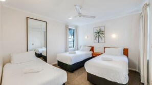 Habitación de hotel con dos camas, ventilador de techo, espejo grande, ventana con cortinas y luz empotrada en la pared.