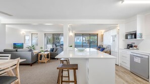 Una cocina moderna con una isla central, un comedor con taburetes altos y una sala de estar con sofá y televisor.