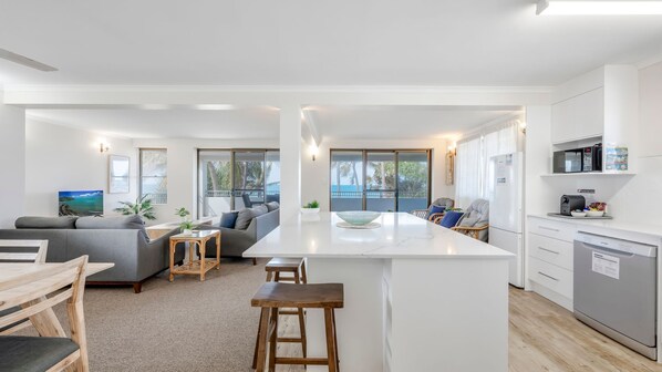 A modern kitchen with a central island, a dining area with bar stools, and a living room with a sofa and a TV. - Horizons 3-16 Miller Bargara (BARGARA)