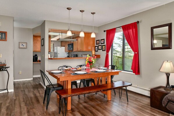 A dining area with a wooden table, chairs, and a bench. There is a kitchen in the background with wooden cabinets and a microwave.