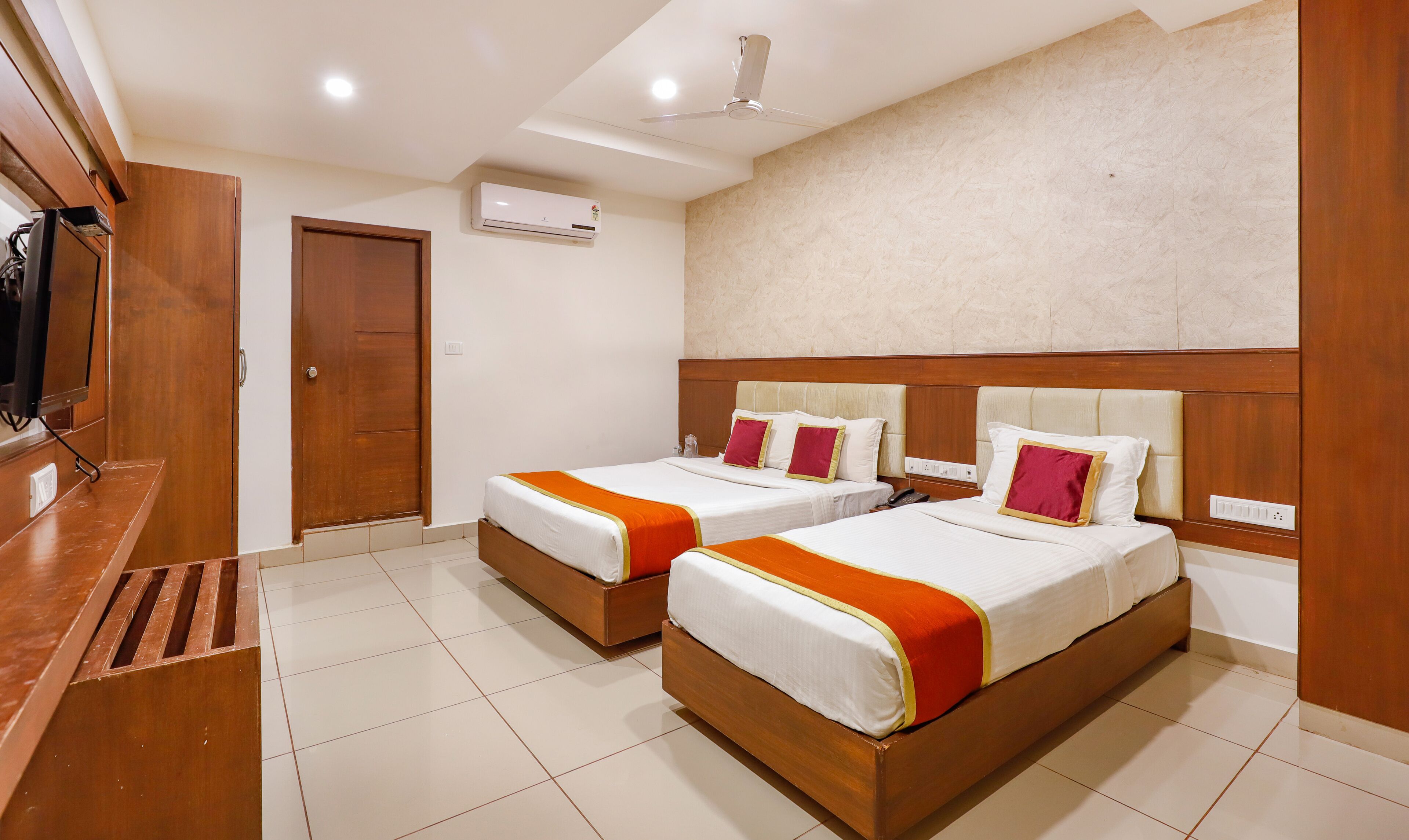A hotel room with two beds, a wooden desk, an air conditioning unit, and a ceiling fan.