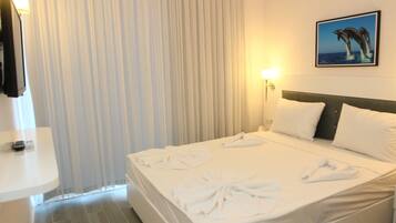 Habitación de hotel con cama, ropa de cama blanca, cabecera, mesita de noche, lámpara, un cuadro en la pared y una ventana con cortinas.