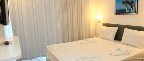 Habitación de hotel con cama, ropa de cama blanca, cabecera, mesita de noche, lámpara, un cuadro en la pared y una ventana con cortinas.