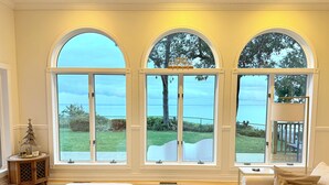 Habitación con tres grandes ventanales de arco que dan a un área verde y al mar. Hay un sofá blanco y un pequeño mueble de madera con un adorno de estrella.