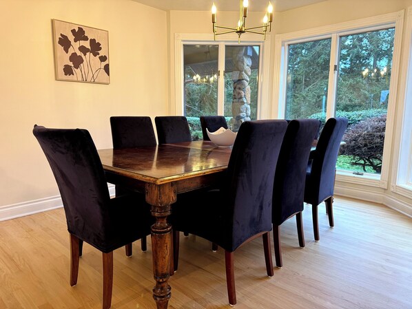 A dining room with a wooden table surrounded by six chairs, large windows with a view of greenery, and a chandelier.