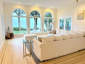 A spacious living room with a large white sofa, a glass coffee table, and a wooden floor. There are three large arched windows providing a view of the outdoors.