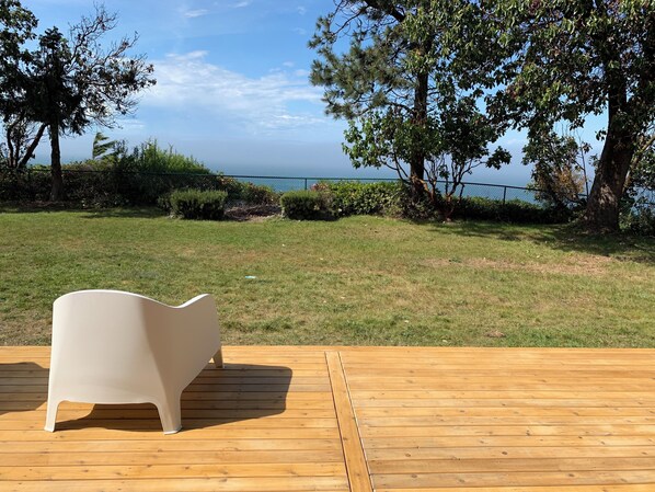 A wooden deck with a white chair, overlooking a grassy area with trees and a view of the sea.