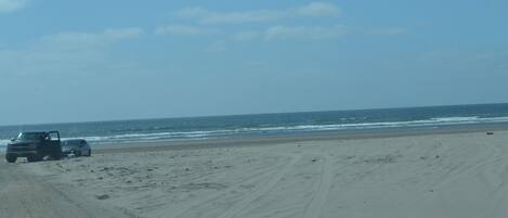 A beach with a vehicle parked on the sand, a clear sky, and the ocean in the background.