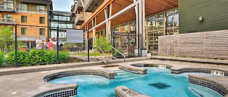 Zona de piscina con varios jacuzzis, rodeada de edificios con balcones y ventanas amplias.