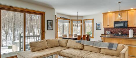 A modern living room with a beige sectional sofa, a glass coffee table, and a view of a deck with snow outside.