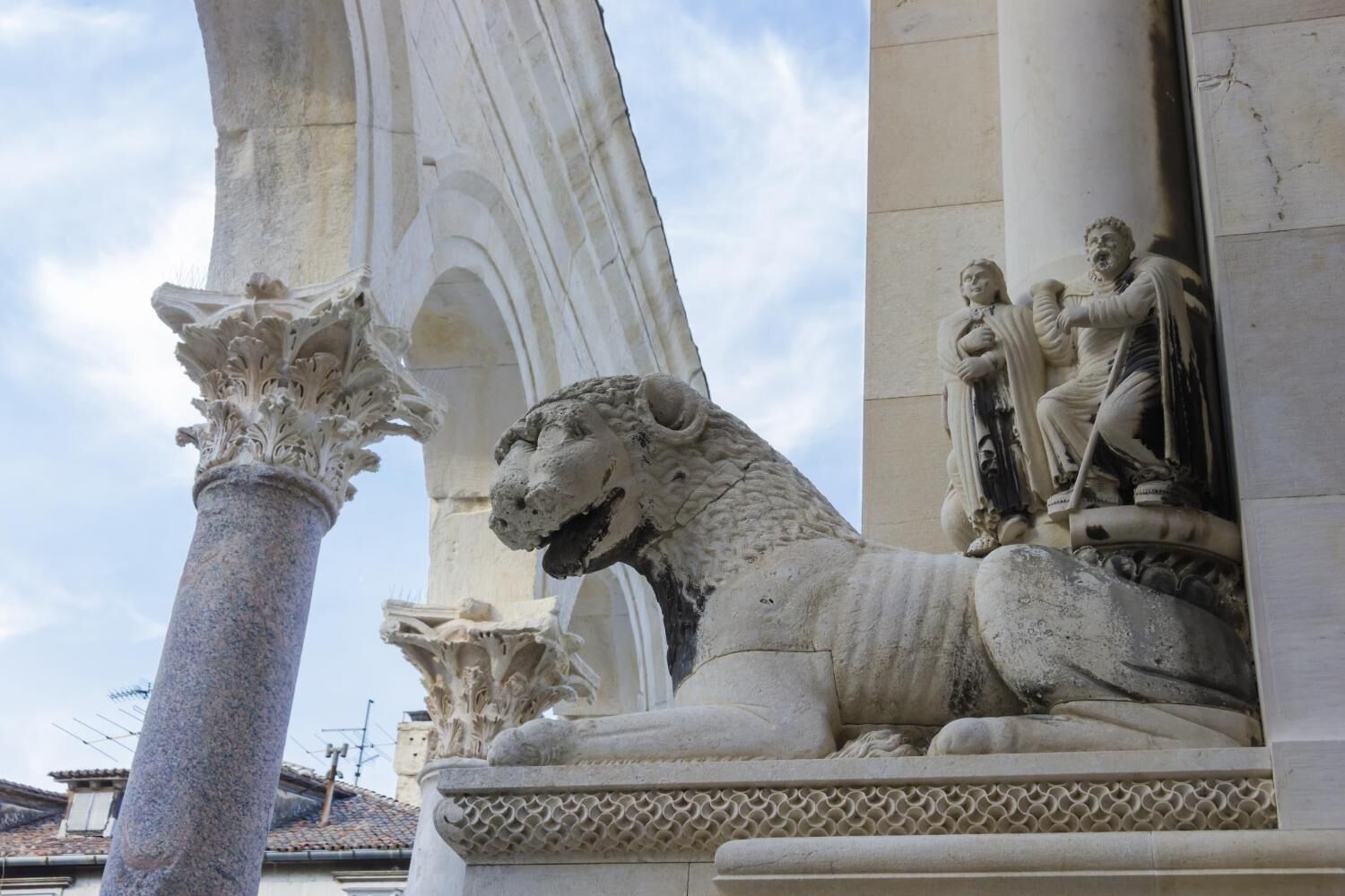 Una escultura de león de piedra con detalles intrincados, ubicada junto a una columna con capiteles ornamentados.
