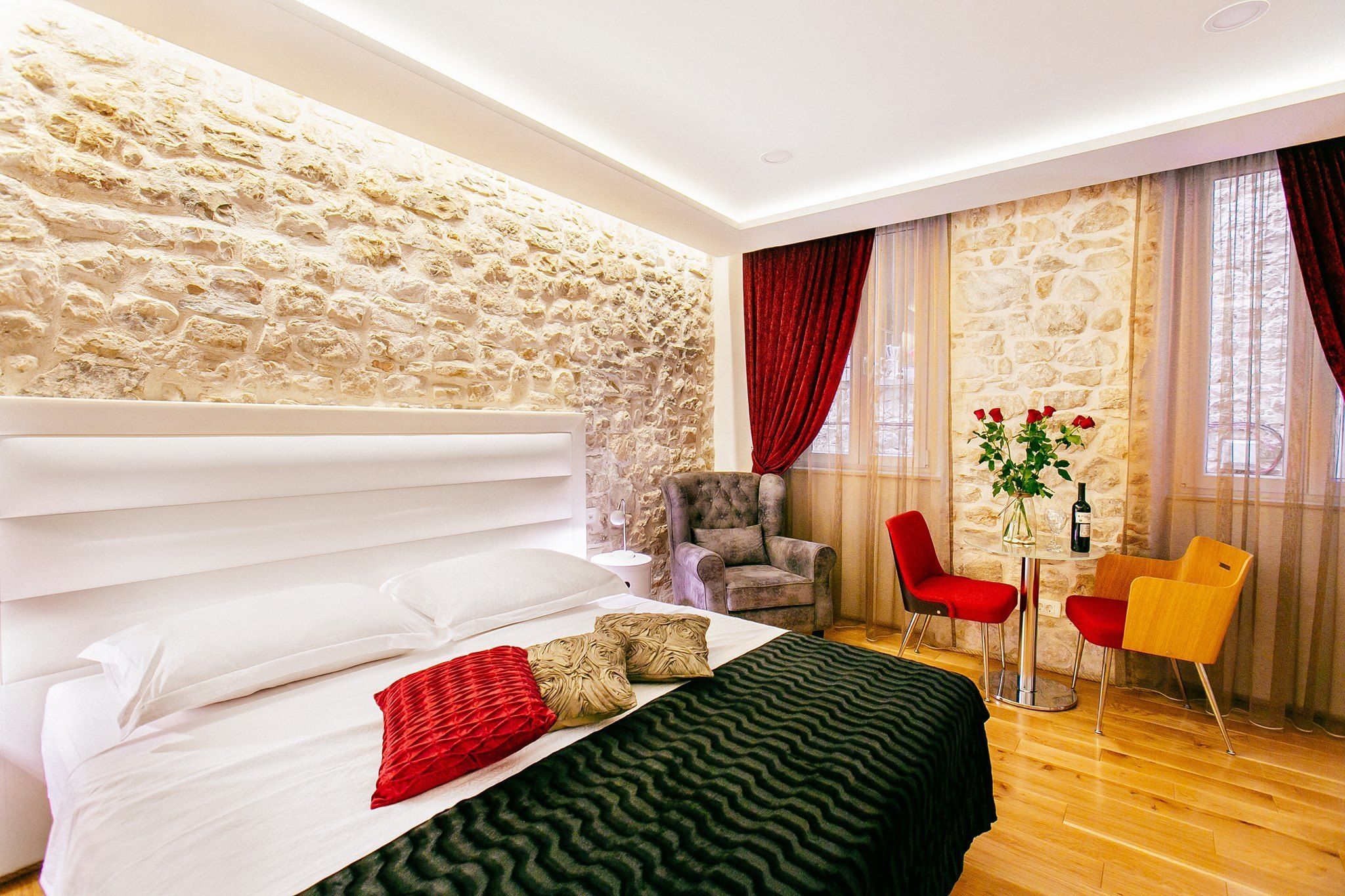 Une chambre avec un mur en pierre, un lit avec une literie blanche, un oreiller rouge, une couverture verte, un fauteuil gris et une table avec des chaises.