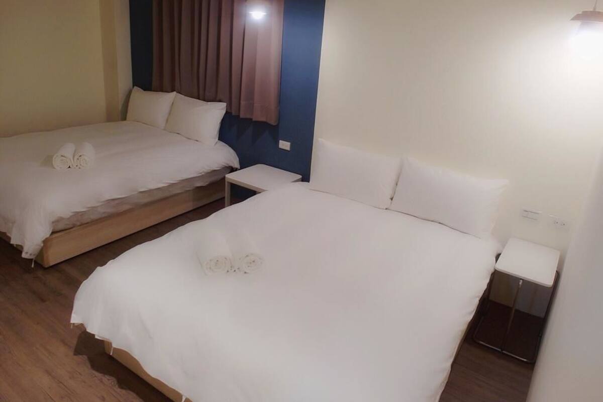 Two single beds with white linens and pillows, a wooden floor, a nightstand with a lamp, and a window with curtains.