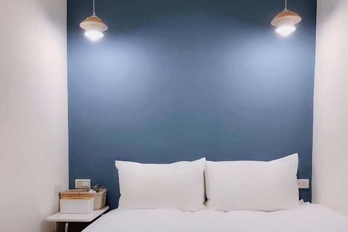 A neatly made bed with white linens in a room with a blue accent wall and two pendant lights.