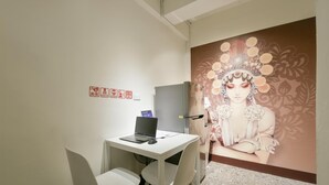 Un pequeño espacio de oficina con un escritorio blanco, dos sillas blancas, una computadora portátil y un refrigerador. En la pared hay un mural que representa a una mujer con adornos florales.