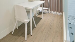 Una habitación pequeña y luminosa con un escritorio y una silla blancos, una lámpara de pie y una ventana con cortinas.