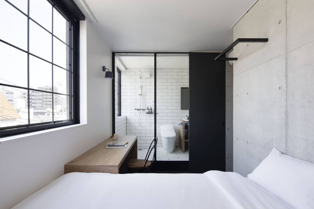A modern hotel room with a large window, a wooden desk, a bed, and a separate bathroom.