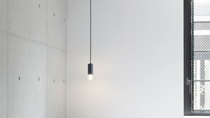 Et minimalistisk hotelværelse med et træskrivebord, en enkelt stol, en seng med hvide sengetæpper og en hængende lampe.