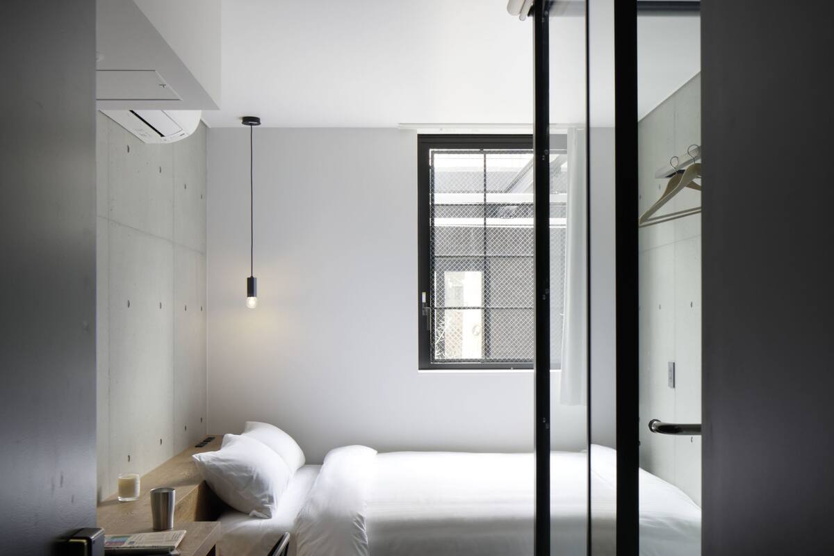 Quarto de hotel pequeno, com uma cama, uma escrivaninha com cadeira, uma mesinha e uma janela que traz luz natural.