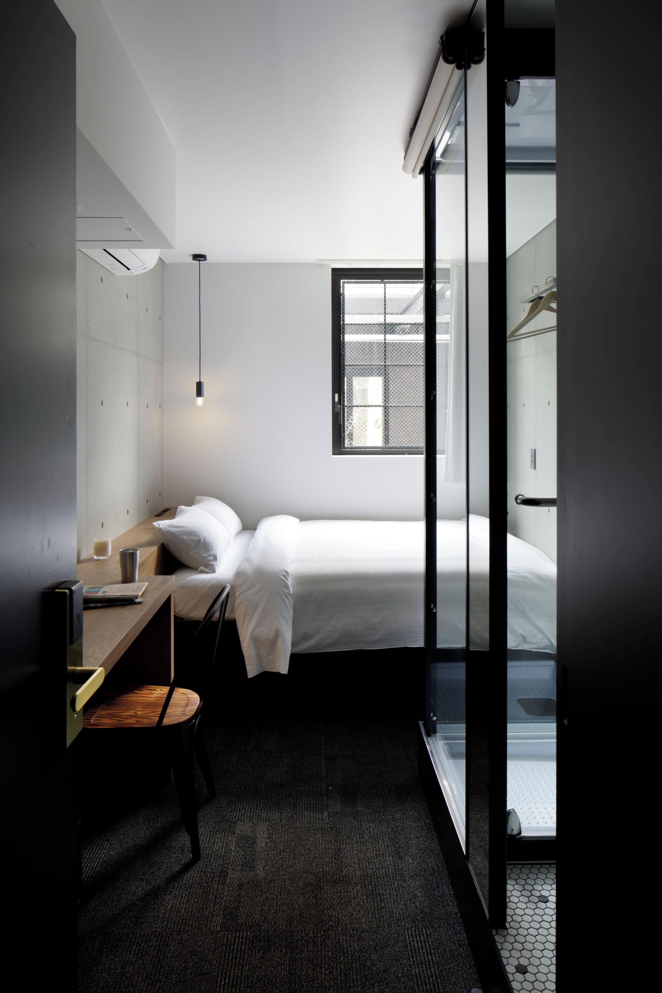A compact hotel room with a bed, a desk, a chair, a small table, a window, and a door leading to another room.