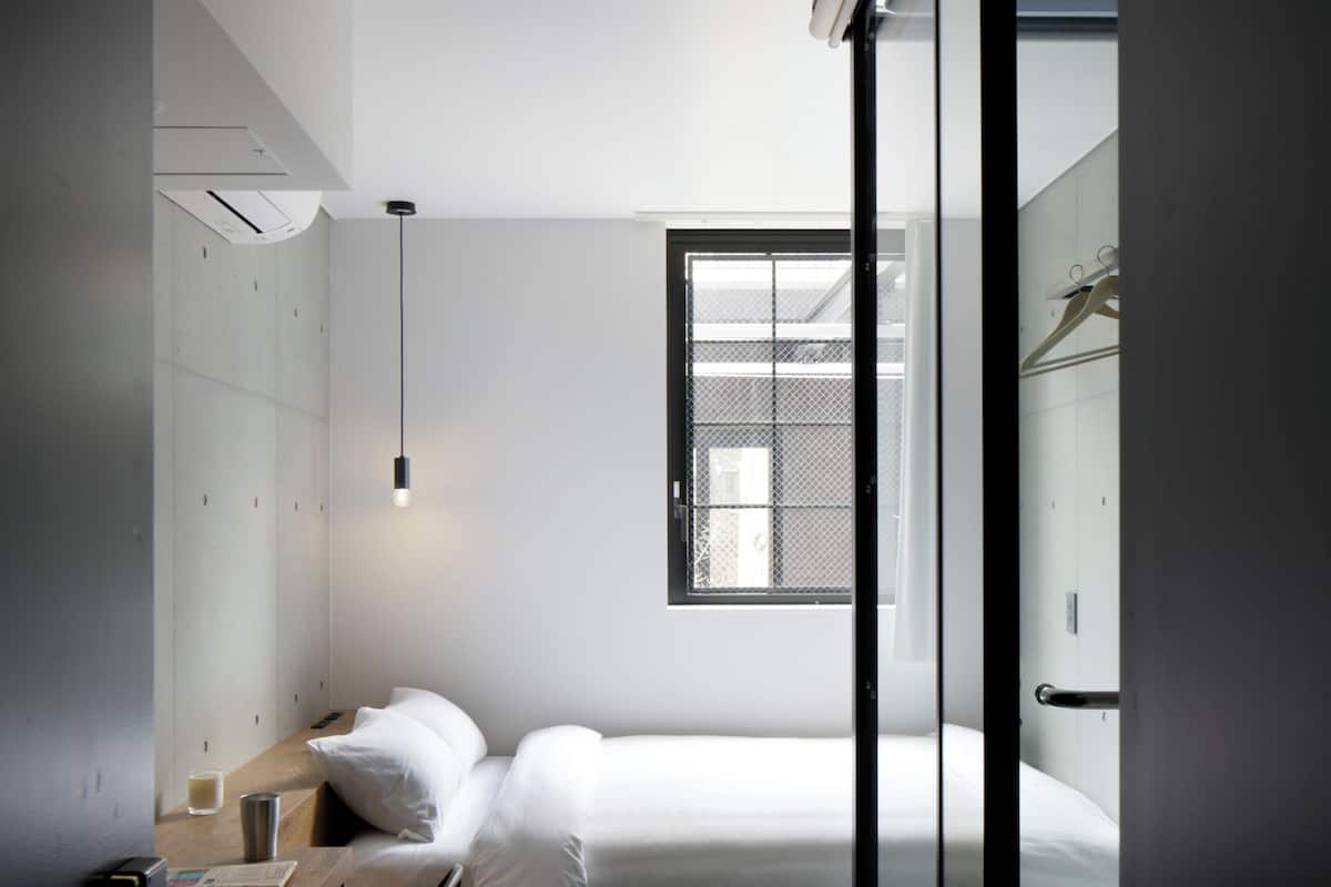 Quarto de hotel pequeno, com uma cama, uma escrivaninha com cadeira, uma mesinha e uma janela que traz luz natural.