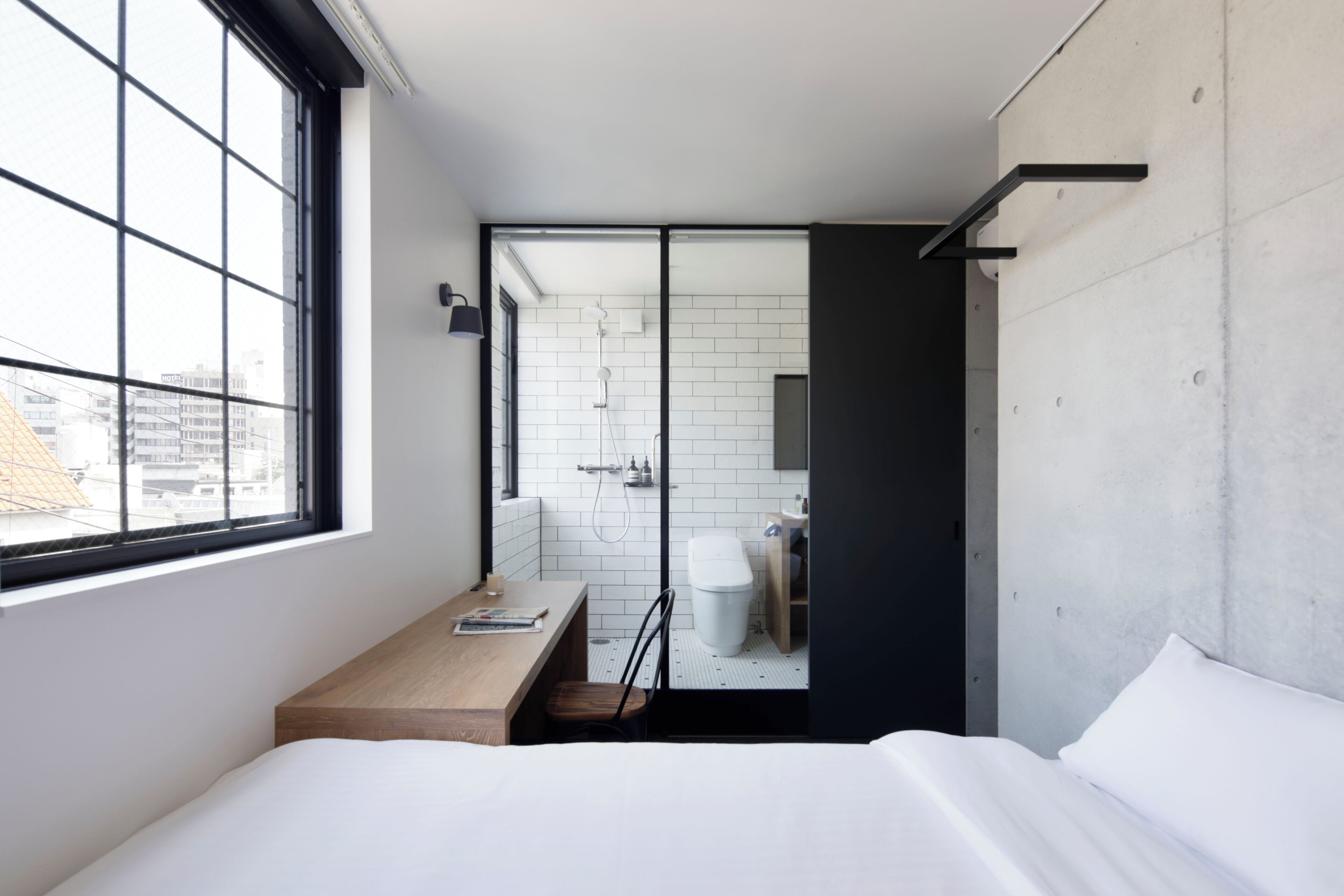 A modern hotel room with a large window, a wooden desk, a bed, and a bathroom with a white tiled wall and a shower.