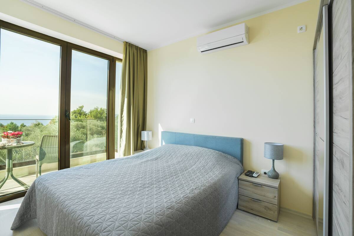 A bedroom with a large bed, a nightstand, a lamp, and a view of greenery through a balcony door.