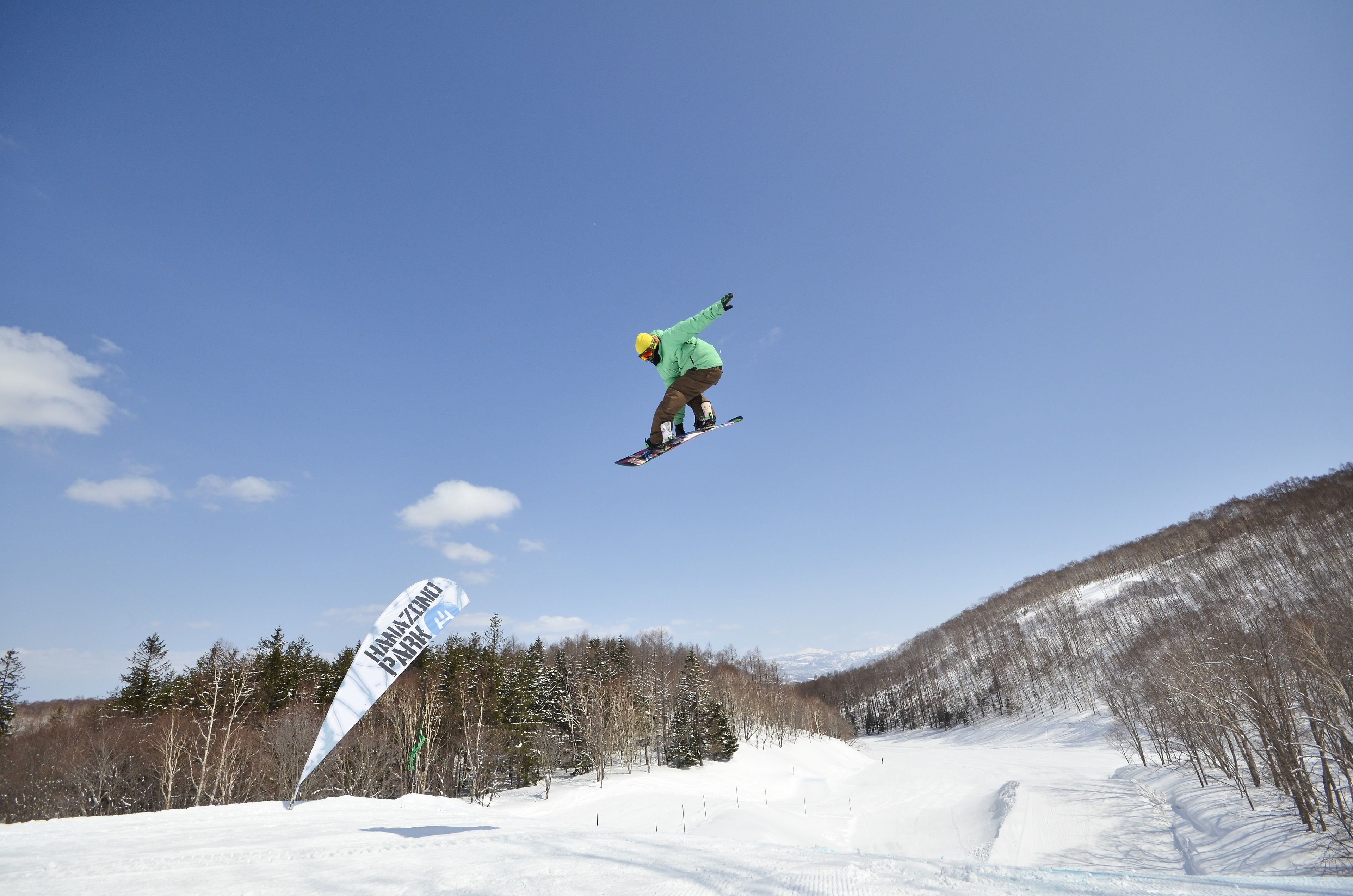 A snowboarder in mid-air performing a jump with a clear blue sky and a snowy landscape in the background.