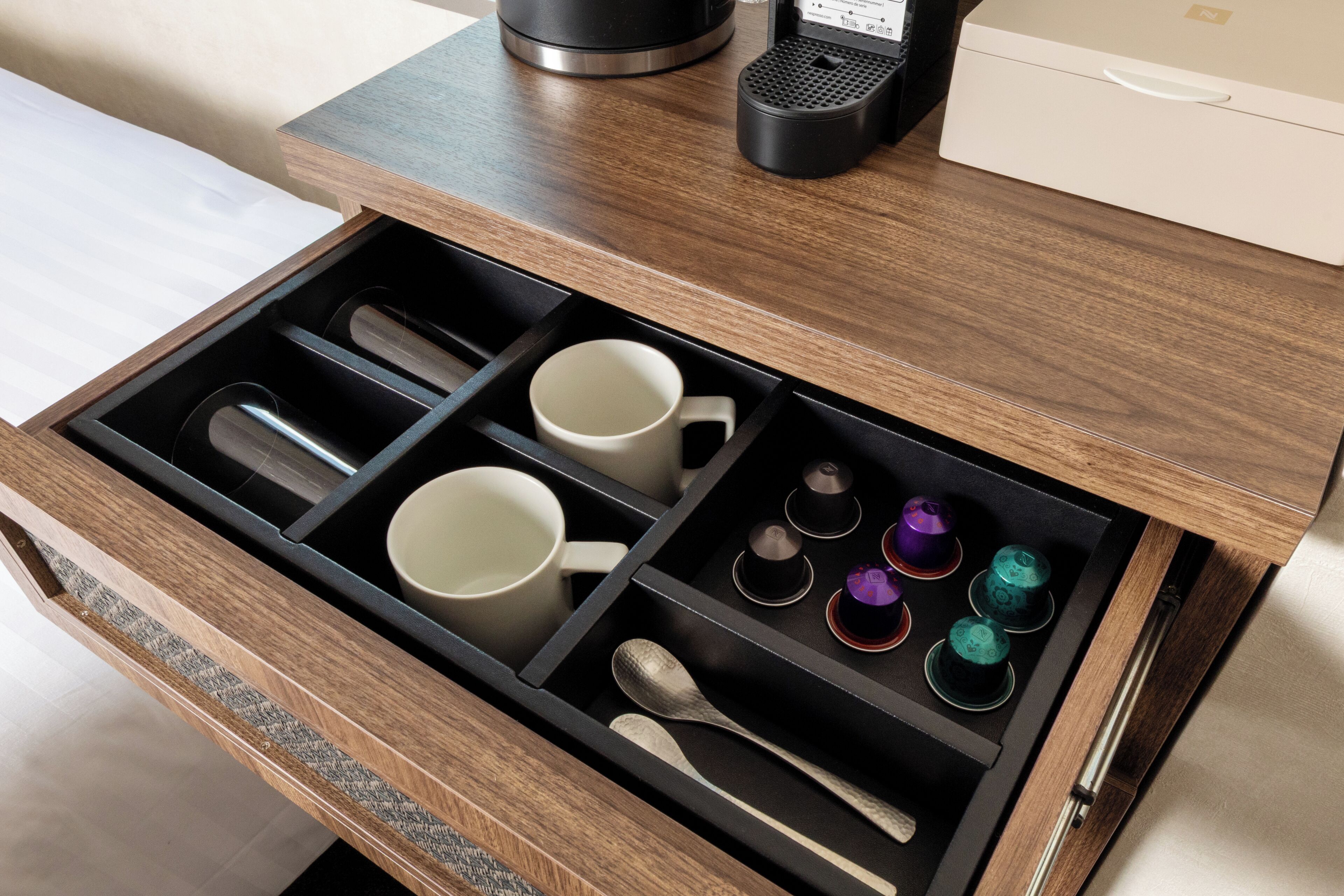 A drawer with compartments containing mugs, a spoon, and a coffee maker.
