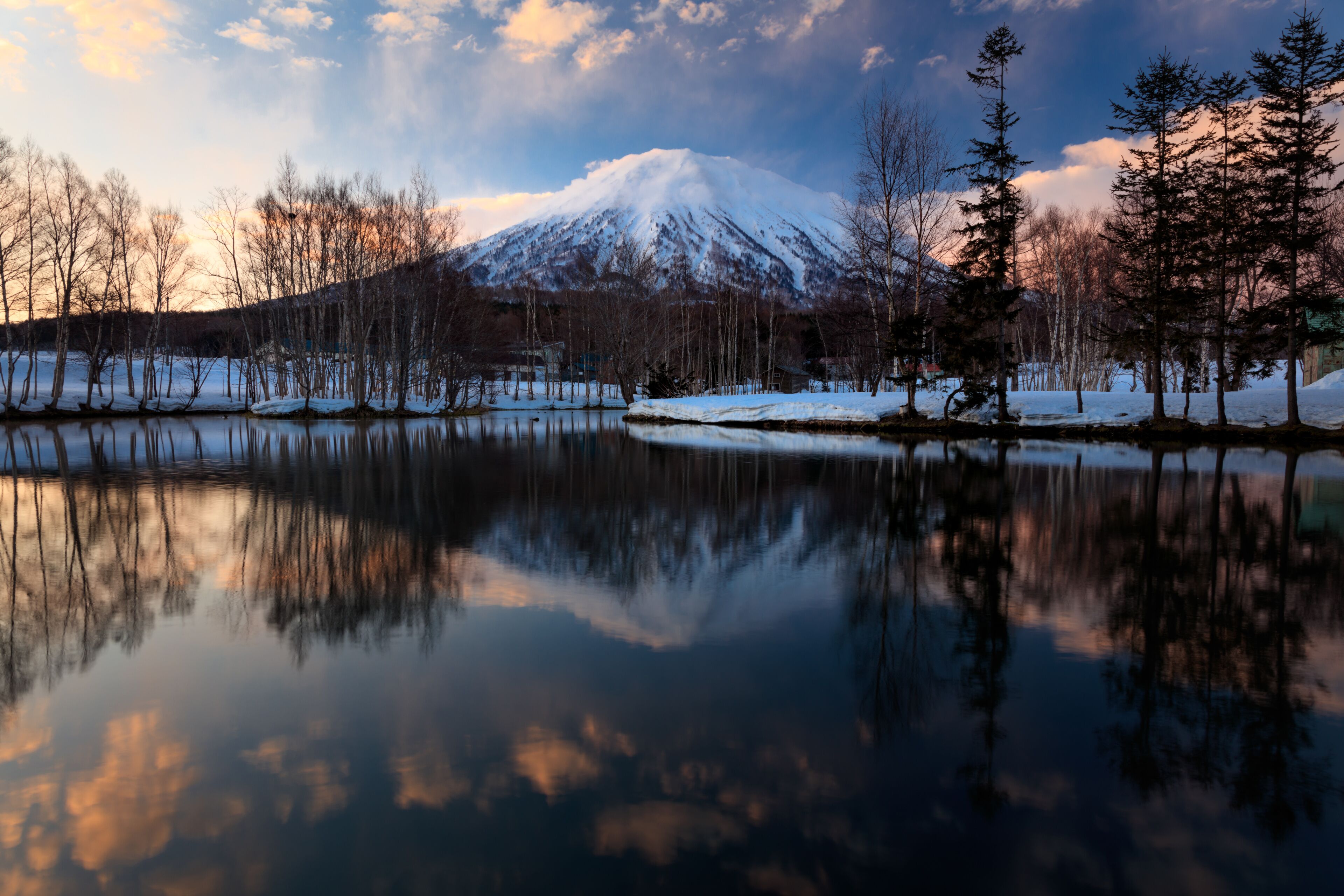A snow-covered mountain, a calm lake, and leafless trees.